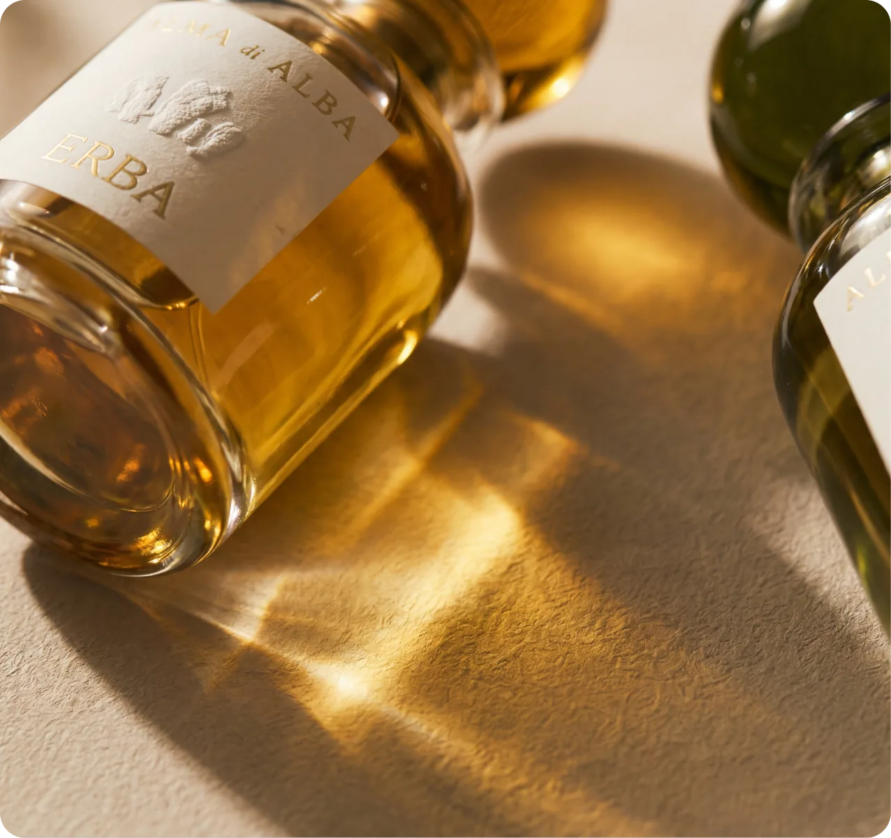 Alma di Alba — Erba bottle close-up laid on its side with an amber tail of sunlight across cream