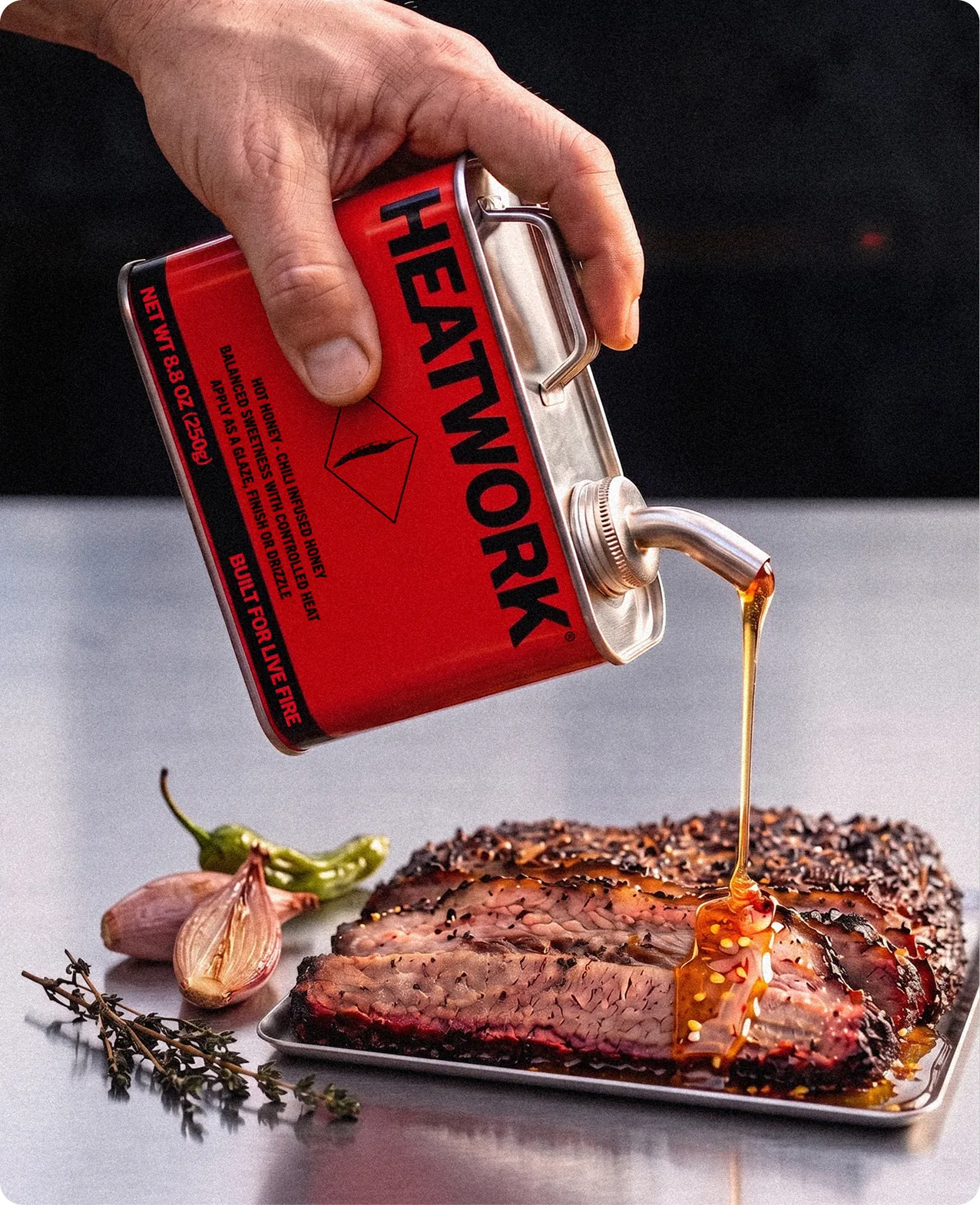 Heatwork: a hand tipping the red tin to pour honey over a sliced, dark-crusted brisket on a steel tray, with a shallot, a green chilli and sprigs of thyme laid beside it