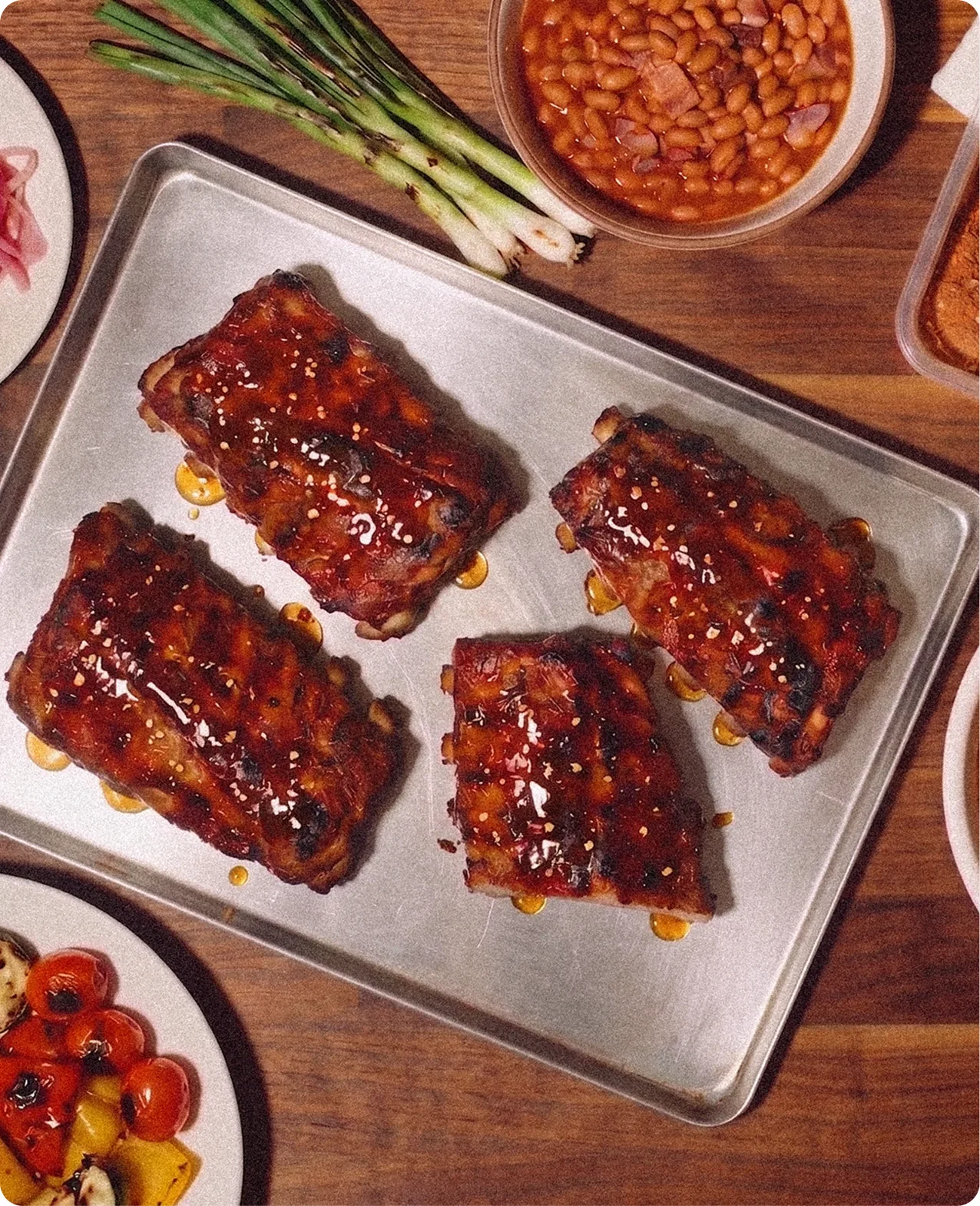 Heatwork: an overhead shot of four glazed rib racks on a steel tray on a wooden table, surrounded by spring onions, a bowl of beans and plates of charred vegetables