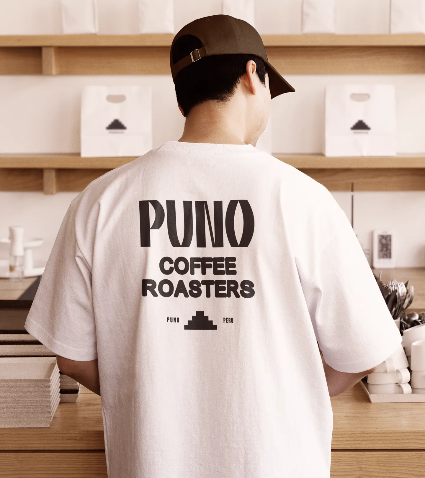 Puno — person from behind wearing a white PUNO COFFEE ROASTERS t-shirt with the pyramid mark, standing in a coffee shop with paper takeaway bags on the shelves behind