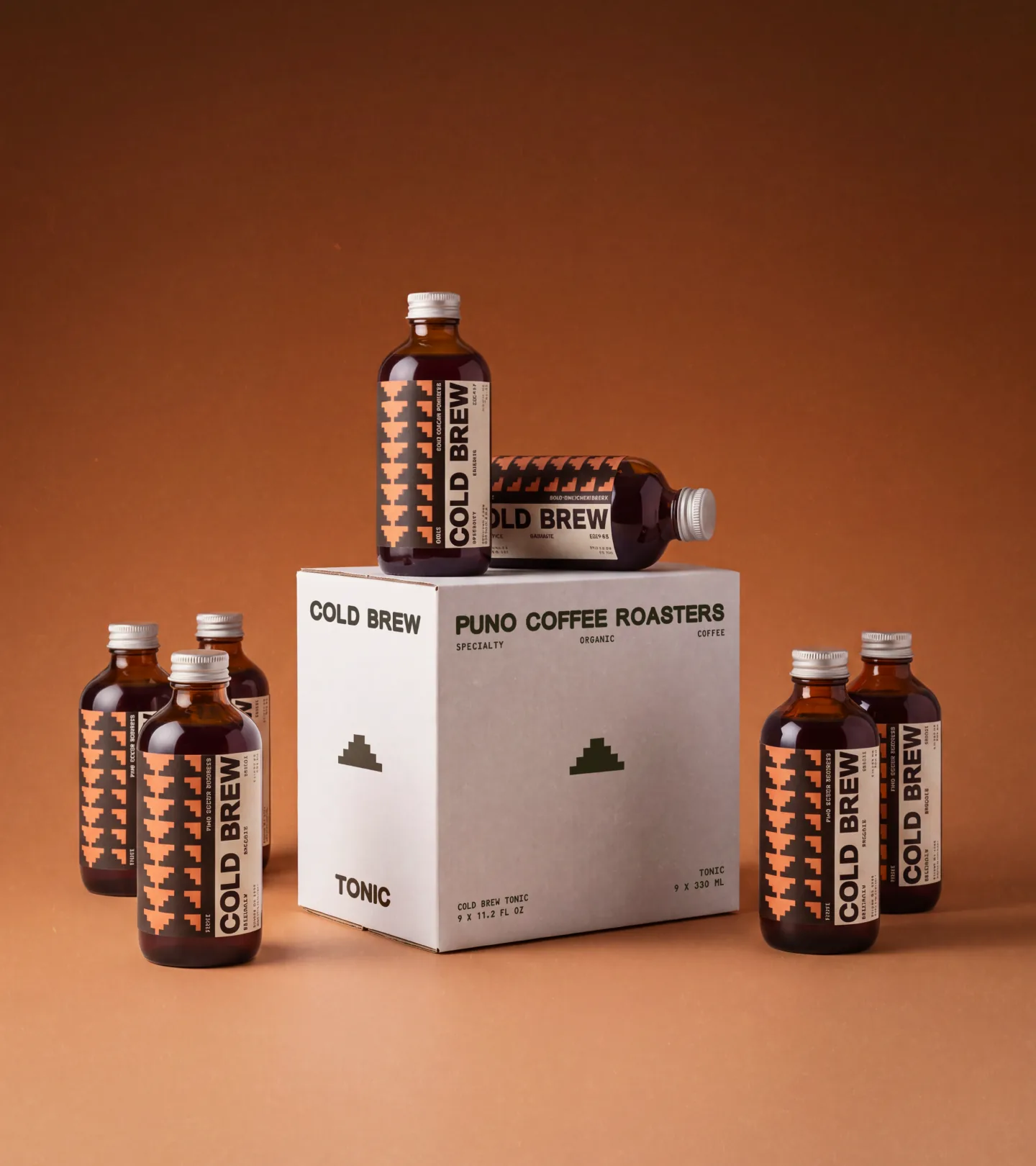 Puno — five amber Cold Brew bottles with peach-and-black pyramid-motif labels arranged around a white 'COLD BREW / PUNO COFFEE ROASTERS' tonic box on a warm terracotta backdrop