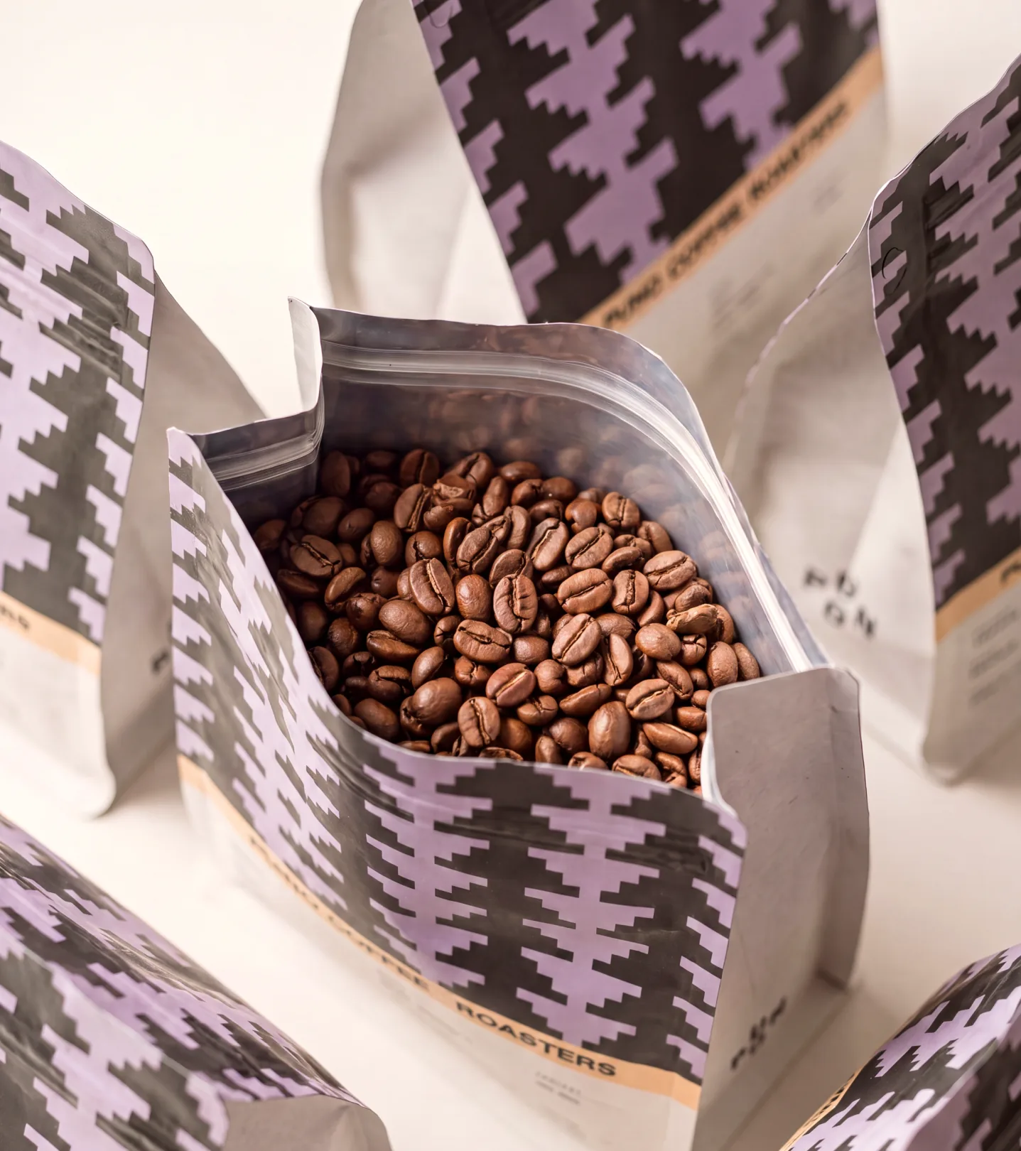 Puno — a cluster of lavender-and-black pyramid-patterned Puno coffee bags standing around a central bag opened at the top to reveal dark-roasted whole beans inside, shot on a cream backdrop
