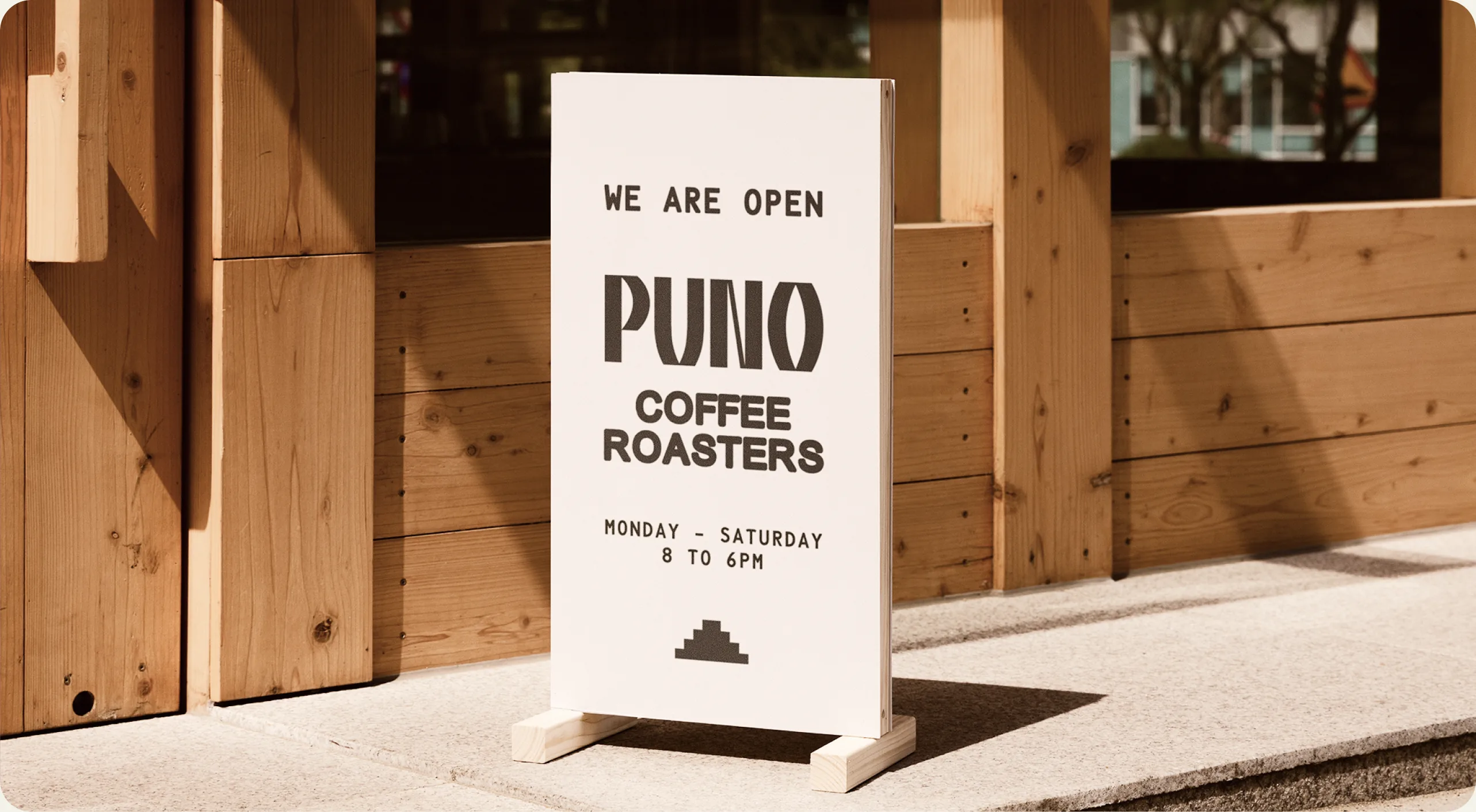 Puno — outdoor A-frame sandwich board in front of the shop reading 'WE ARE OPEN / PUNO COFFEE ROASTERS / MONDAY – SATURDAY / 8 TO 6PM' with the pyramid mark, standing on a stone step against a warm wooden slat facade