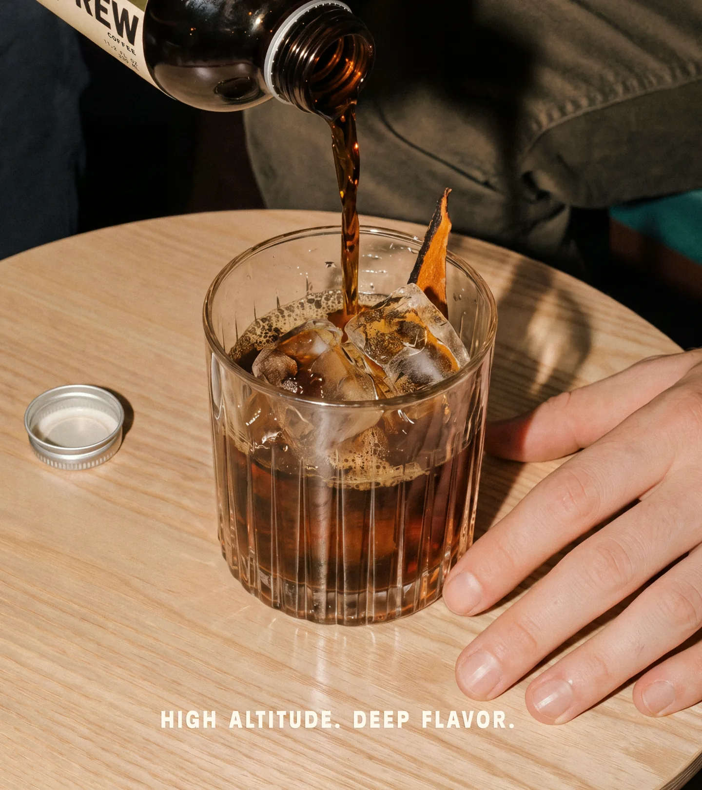 Puno — hand pouring cold brew from an amber Puno bottle over ice and a dehydrated orange slice into a fluted rocks glass on a light wood table, with 'HIGH ALTITUDE. DEEP FLAVOR.' set at the base of the frame