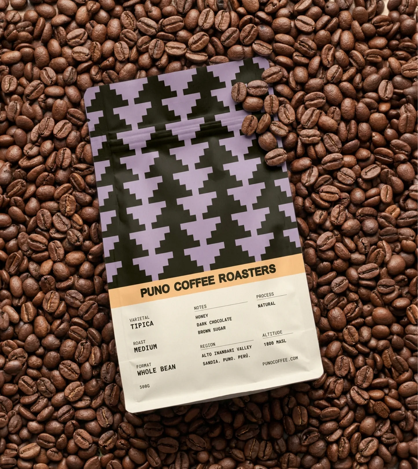 Puno — a lavender-and-black pyramid-patterned Puno whole-bean coffee bag lying flat on a bed of dark-roasted beans, with the full 'PUNO COFFEE ROASTERS' spec label detailing the Tipica varietal, medium roast, honey / dark chocolate / brown sugar tasting notes, natural process, and Alto Inanbari Valley origin in Sandia, Puno, Perú at 1800 masl