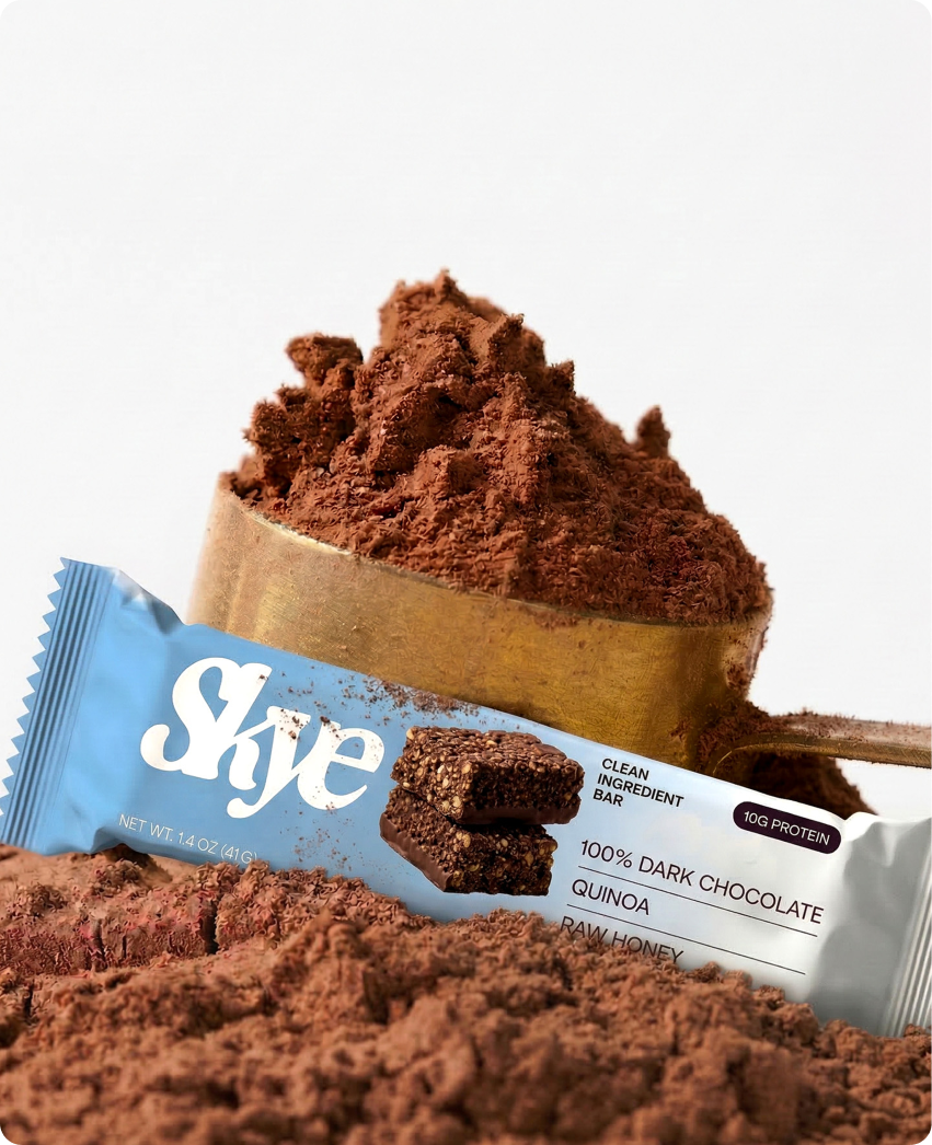 Skye — a dark chocolate and quinoa protein bar leaning against a scoop of raw cacao powder on a clean neutral backdrop