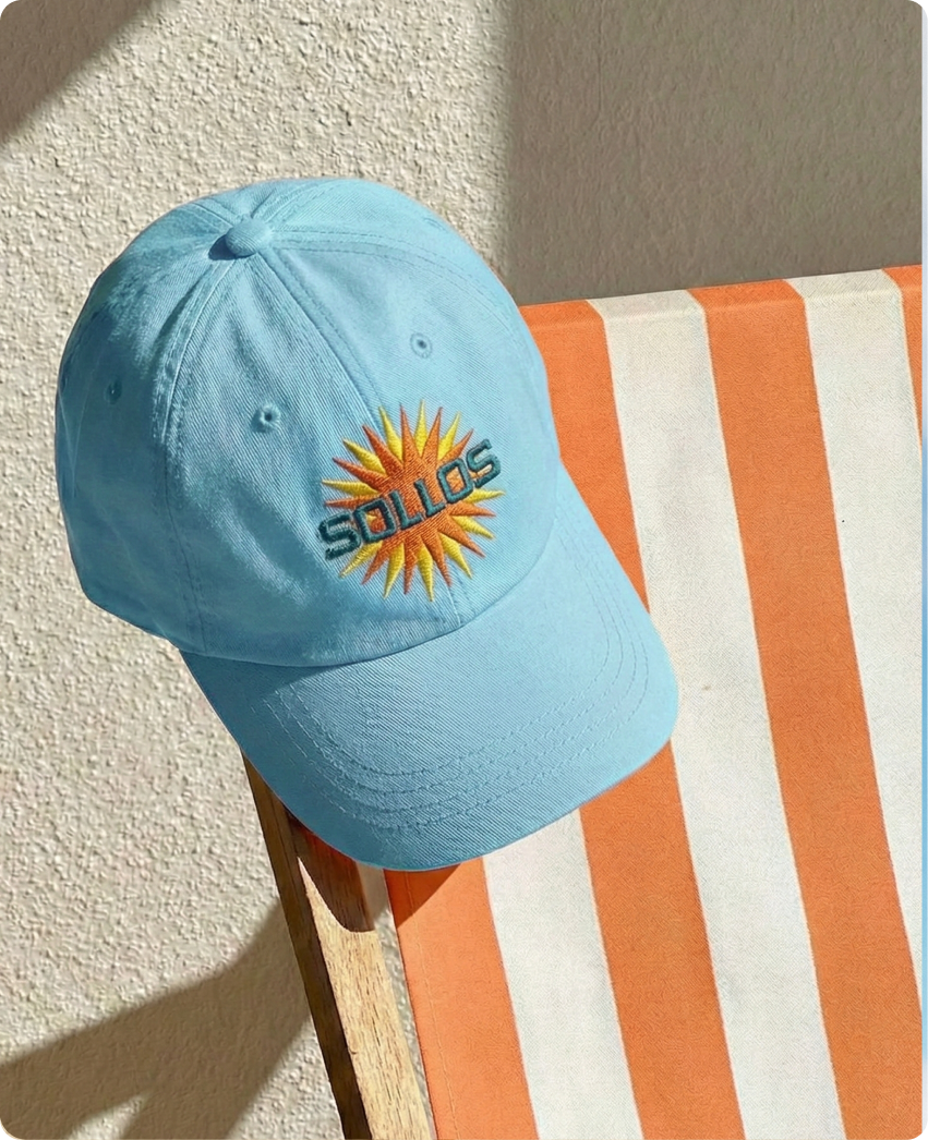 Sollos — a sky-blue cap embroidered with the Sollos sunburst logo resting on an orange-and-white striped beach chair