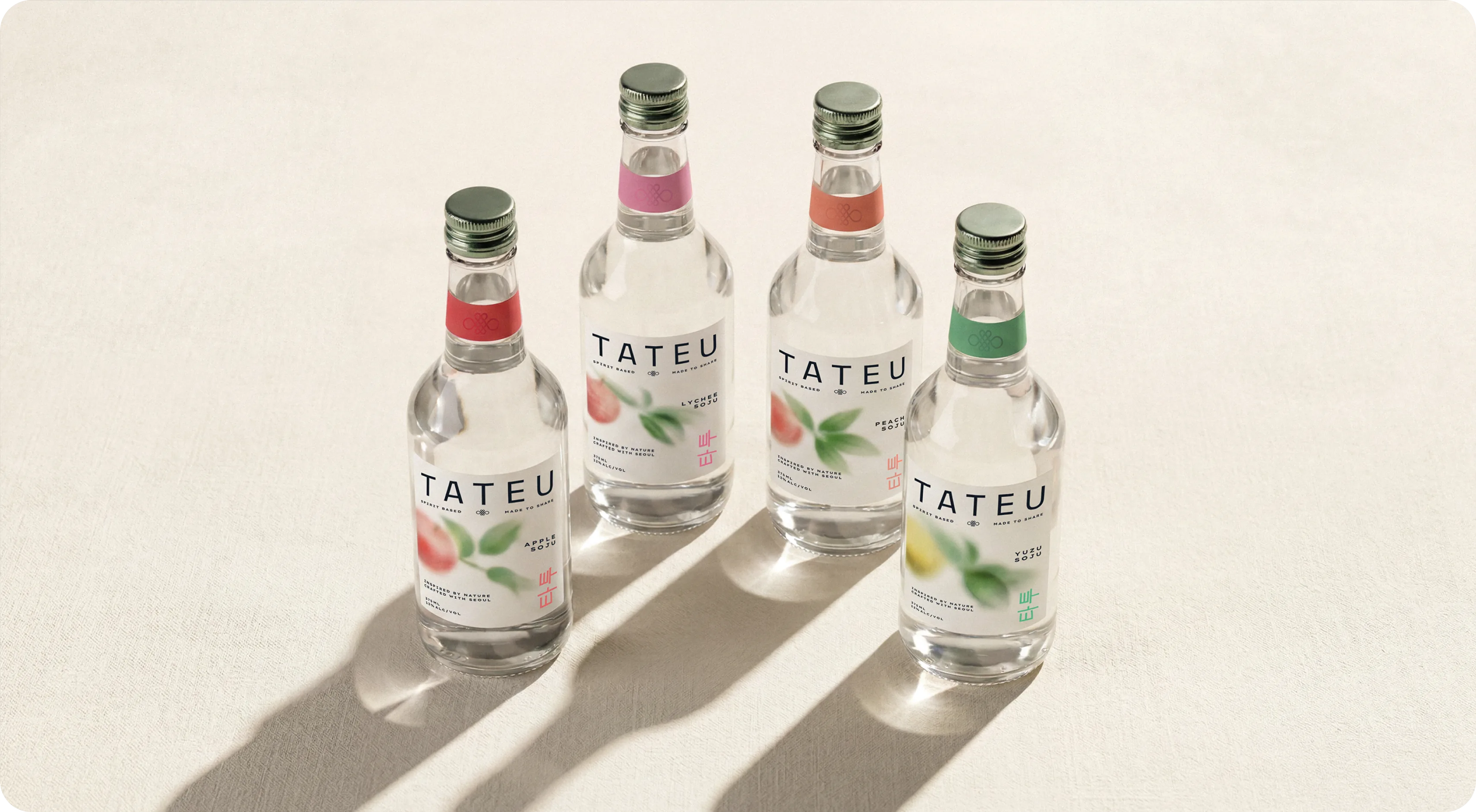 Tateu — the four-bottle lineup standing in a row on a cream surface in raking light, each 375ml bottle carrying the TATEU wordmark, a soft-focus fruit illustration and a different muted neck band for Peach, Lychee, Apple and Yuzu