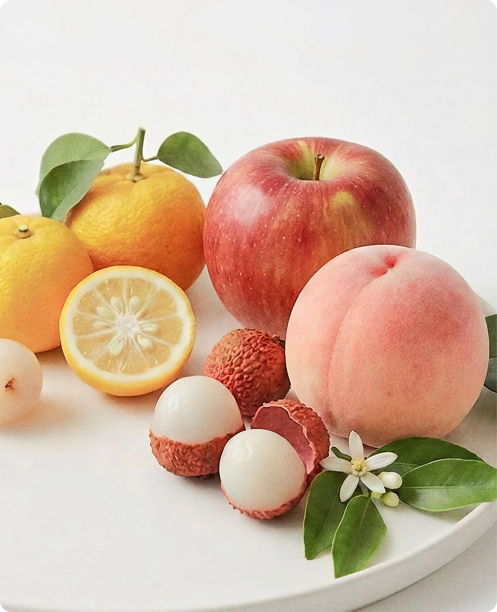 Tateu — the four source fruits arranged as a still life on a white ceramic plate: a whole peach, a red apple, yuzu and a halved lemon, peeled and unpeeled lychees, and an orange blossom with glossy leaves