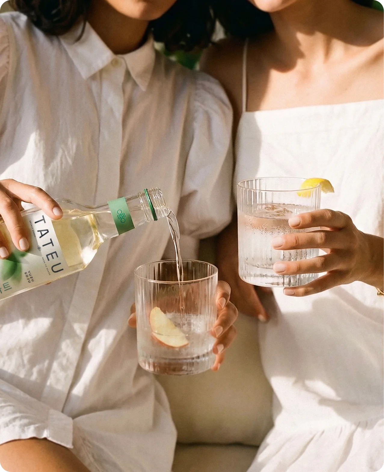 Tateu — two people in white linen sitting together, one pouring soju from a Tateu bottle into a fluted rocks glass garnished with a peach slice while the other holds a filled glass with a lemon twist
