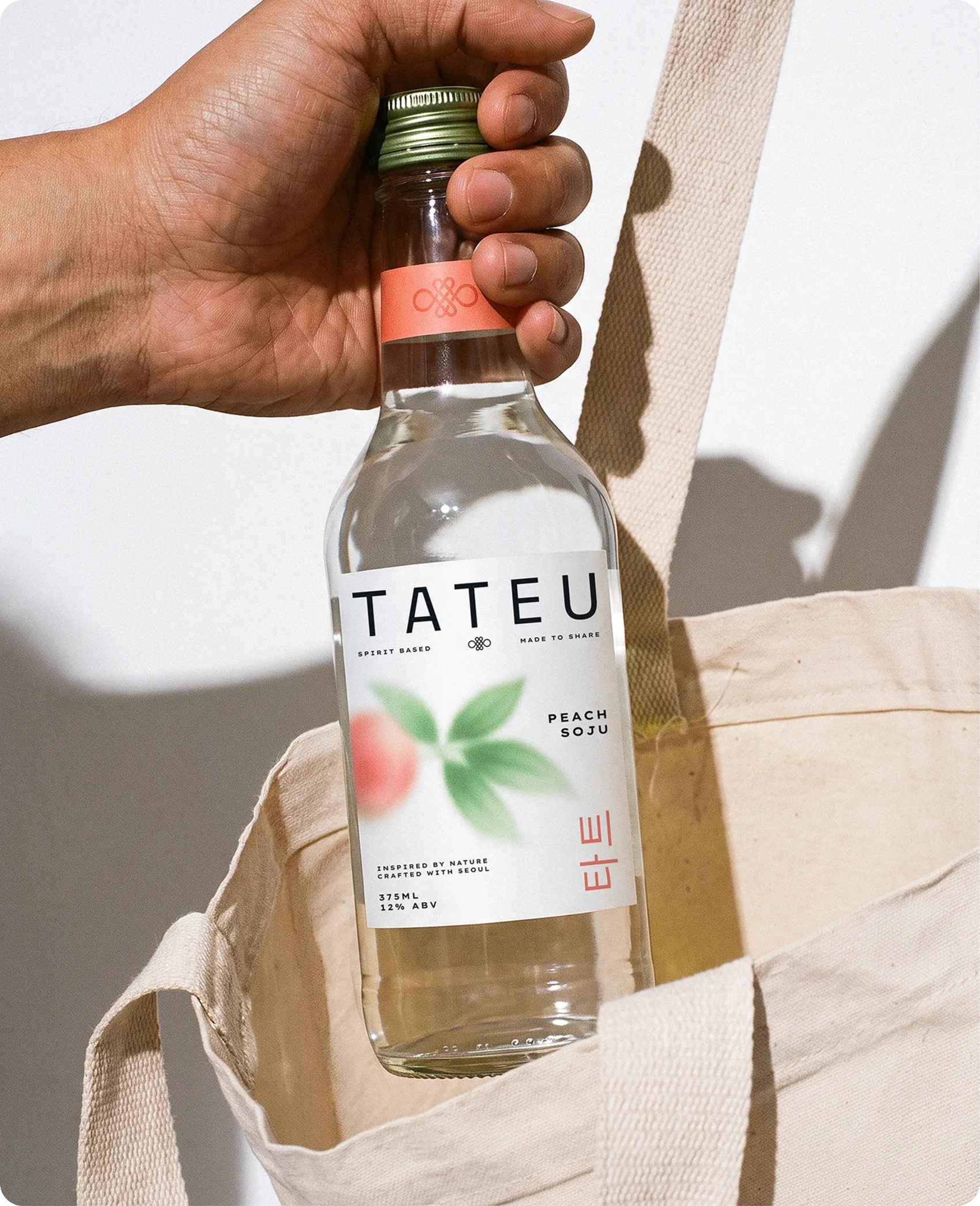 Tateu — a hand lifting a bottle of Peach Soju out of a natural canvas tote bag, the bottle catching hard sunlight against a cream wall