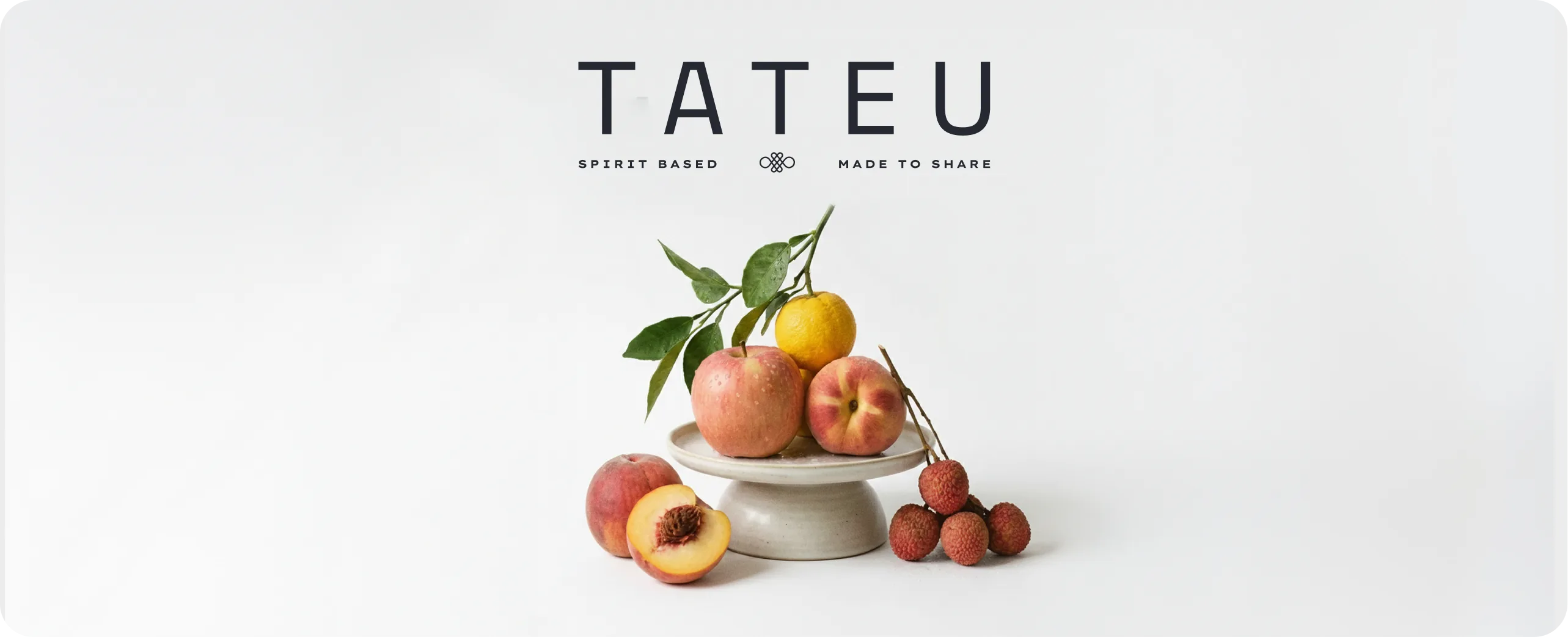 Tateu — brand hero with the widely spaced TATEU wordmark above 'SPIRIT BASED' and 'MADE TO SHARE' either side of the looped monogram, over a ceramic stand holding a peach, an apple, a yuzu and a bunch of lychees on a soft cream backdrop
