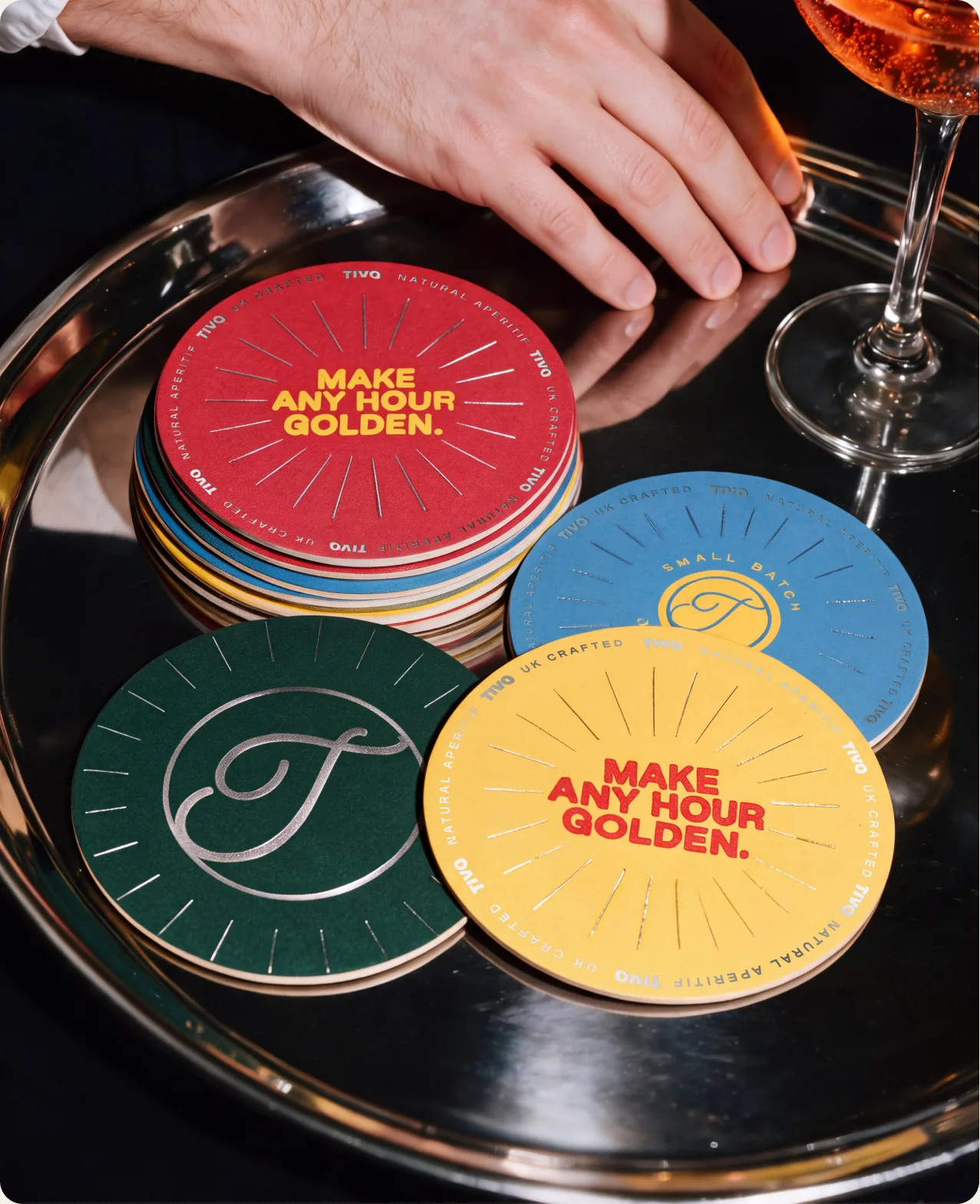 Tivo: a hand reaching across a silver tray of circular coasters, a red and a yellow one reading make any hour golden, a dark green one carrying the cursive T and a blue one reading small batch, beside the stem of a cocktail glass