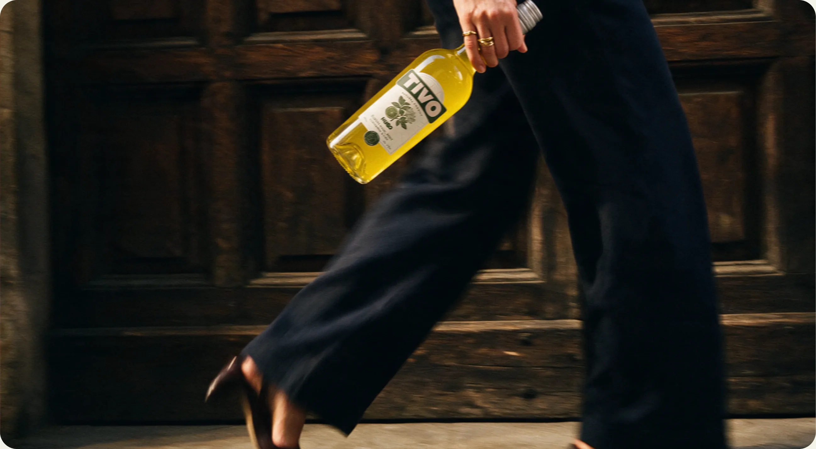 Tivo: a person in wide dark trousers walking past a dark panelled door, carrying a yellow Hugo bottle loosely at their side