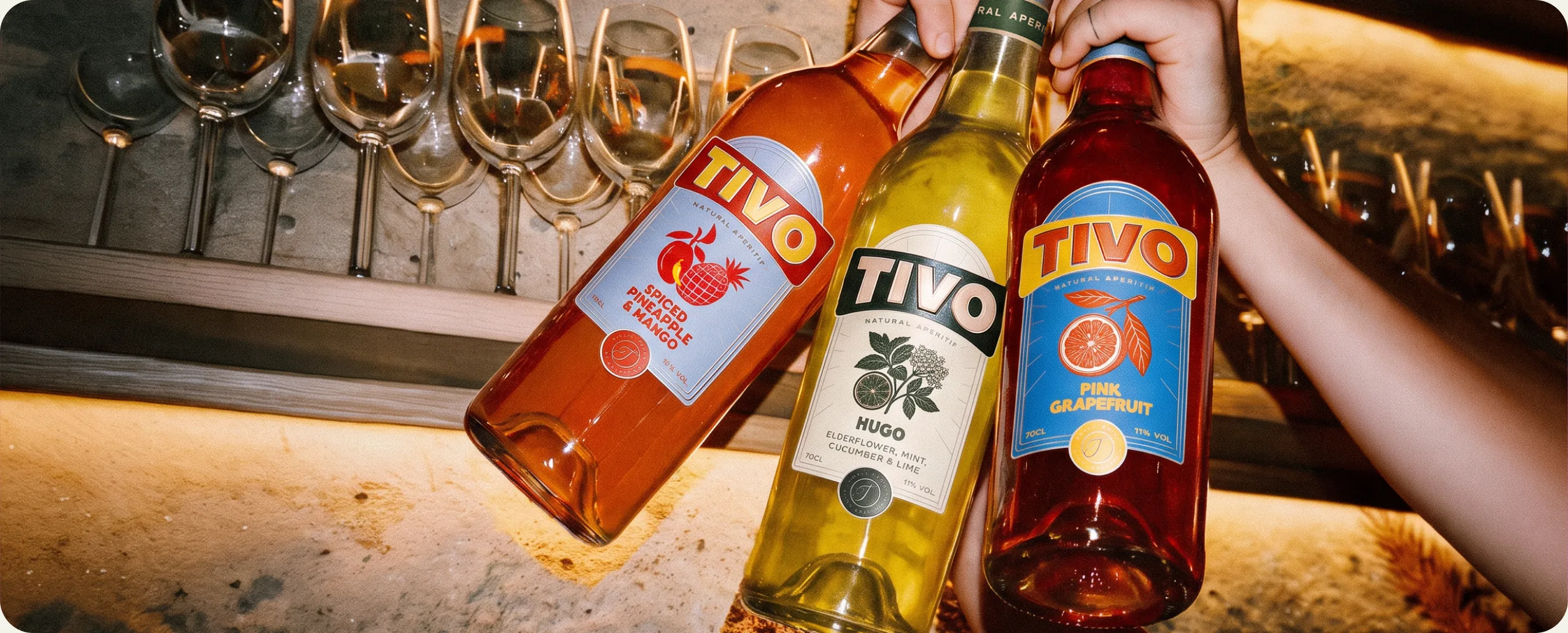 Tivo: a hand setting down three aperitif bottles lying along a bar counter, Spiced Pineapple and Mango in orange, Hugo in yellow green and Pink Grapefruit in red, with a rail of upturned wine glasses behind