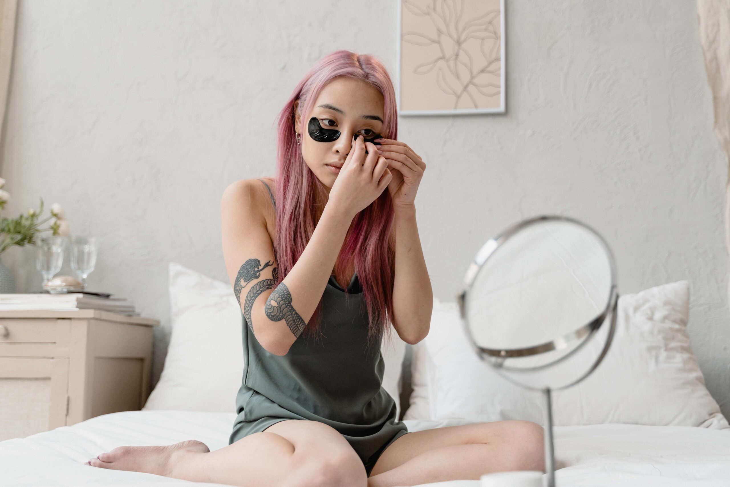 Advertisement of girl sitting in her bed using black under eye pads while she's looking herself ni the mirror